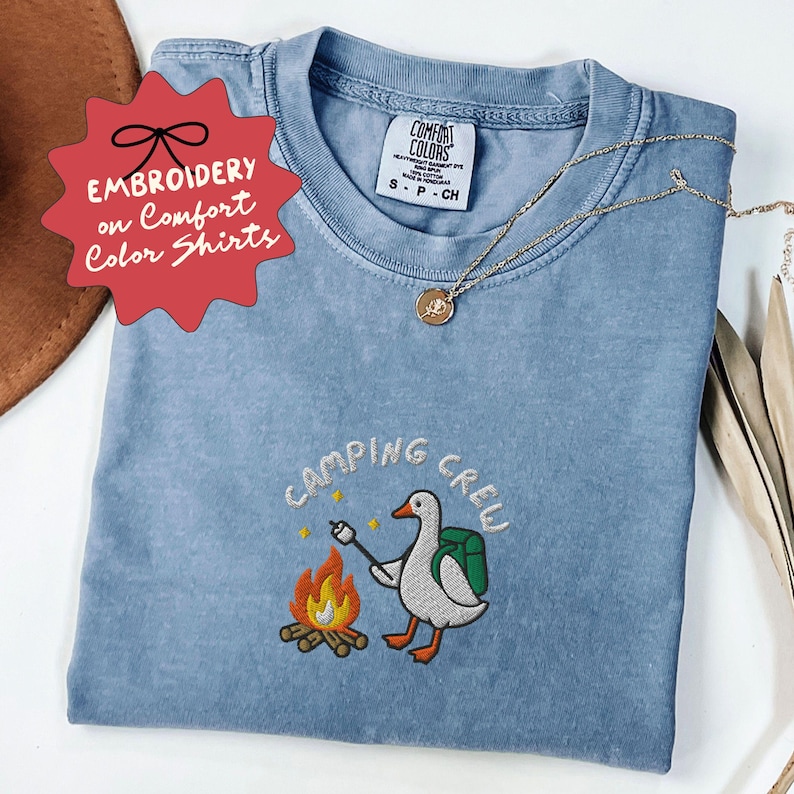 Op de afbeelding: Lichtblauw Comfort Colors T-shirt met een geborduurd ontwerp met een gans die een marshmallow roostert boven een kampvuur. De tekst "CAMPING CREW" staat boven het ontwerp. Het shirt heeft ook de tekst "EMBROIDERY on Comfort Color Shirts" op een rode ster.