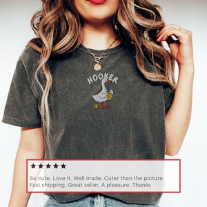 May include: Dark gray t-shirt with the word "HOOKER" above an embroidered goose holding a fishing net. The goose has orange feet and is wearing a yellow hat. The shirt is a casual style.