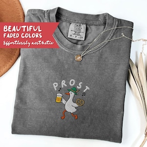 May include: A gray Comfort Colors t-shirt with the word "PROST" embroidered above a cartoon goose wearing a green hat and holding a beer and pretzel. The shirt also has a silver necklace. Text on the shirt reads "BEAUTIFUL FADED COLORS, Effortlessly aesthetic."