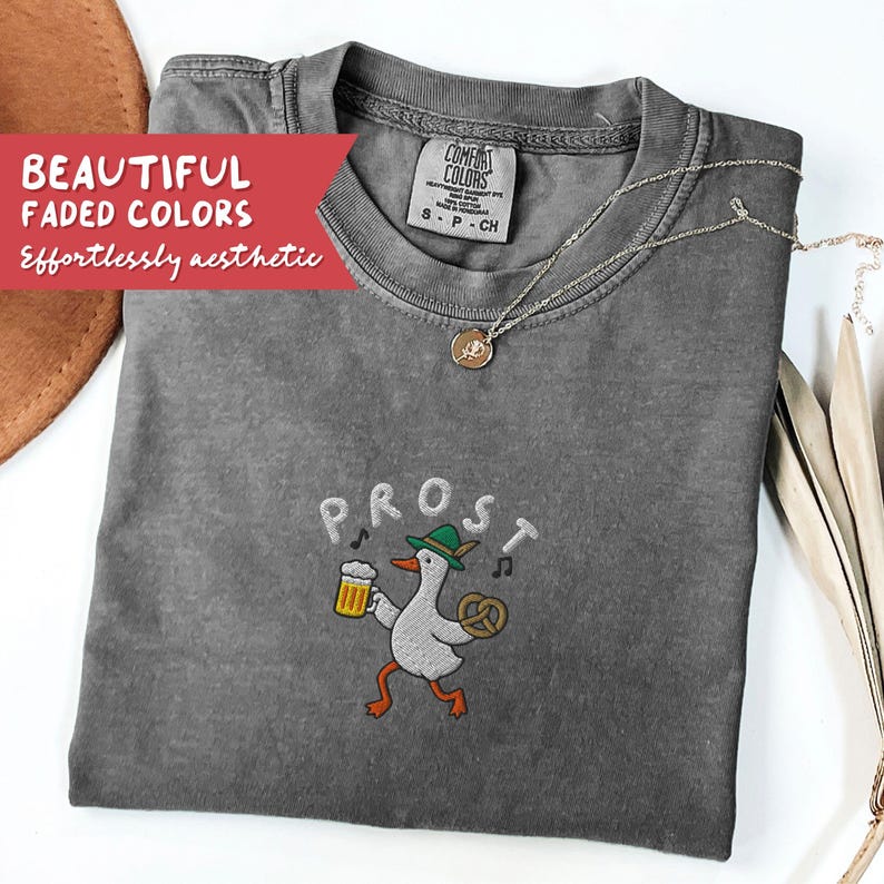 May include: A gray Comfort Colors t-shirt with the word "PROST" embroidered above a cartoon goose wearing a green hat and holding a beer and pretzel. The shirt also has a silver necklace. Text on the shirt reads "BEAUTIFUL FADED COLORS, Effortlessly aesthetic."