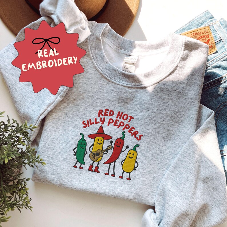 K&ouml;nnte beinhalten: Ein hellgraues Sweatshirt mit dem Text "Red Hot Silly Peppers" und einer Grafik von vier anthropomorphen Chilischoten. Eine Paprika spielt Gitarre. Das Sweatshirt hat ein rotes Stern-Tag mit der Aufschrift "Real Embroidery".