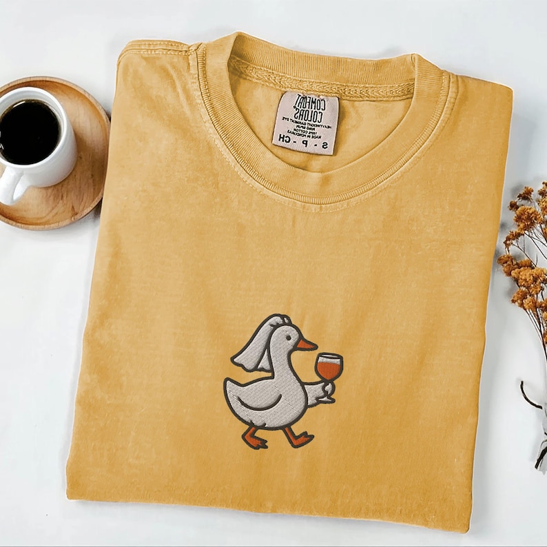 May include: Mustard yellow t-shirt featuring an embroidered cartoon duck wearing a chef's hat and holding a glass of red wine. The shirt has a crew neck and a tag on the collar. A cup of coffee and dried flowers are in the background.