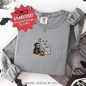 May include: A gray long-sleeve shirt with an embroidered Halloween design. The design features a grim reaper, a ghost roasting marshmallows over a campfire, a crescent moon, and stars. The shirt has the text "EMBROIDERY Original design by seller".