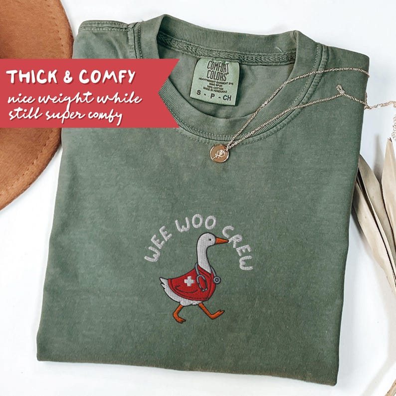 May include: An olive green t-shirt with the embroidered phrase "WEE WOO CREW" and a cartoon goose wearing a red medical coat. A gold necklace with a pendant rests on the shirt. The shirt has the text "THICK & COMFY" on a red banner.