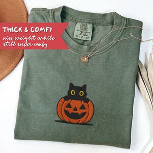 K&ouml;nnte beinhalten: Olivgr&uuml;nes T-Shirt mit einer schwarzen Katze, die aus einem orangefarbenen K&uuml;rbis lugt. Auf dem Shirt steht "THICK & COMFY nice weight while still super comfy" auf einem roten Banner. Eine silberne Halskette mit Anh&auml;nger ist ebenfalls zu sehen.