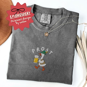 May include: A gray Comfort Colors t-shirt with the embroidered word "PROST" above a cartoon goose wearing a green hat and holding a beer stein. The shirt has a small tag and a silver necklace. The design is labeled "Embroidery Original design by seller."