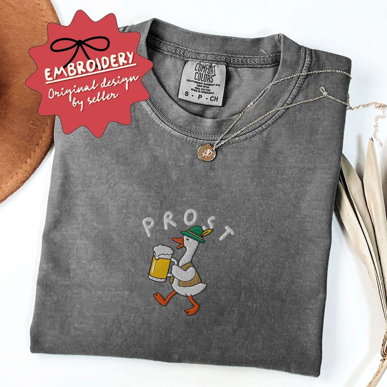 May include: A gray Comfort Colors t-shirt with the embroidered word "PROST" above a cartoon goose wearing a green hat and holding a beer stein. The shirt has a small tag and a silver necklace. The design is labeled "Embroidery Original design by seller."
