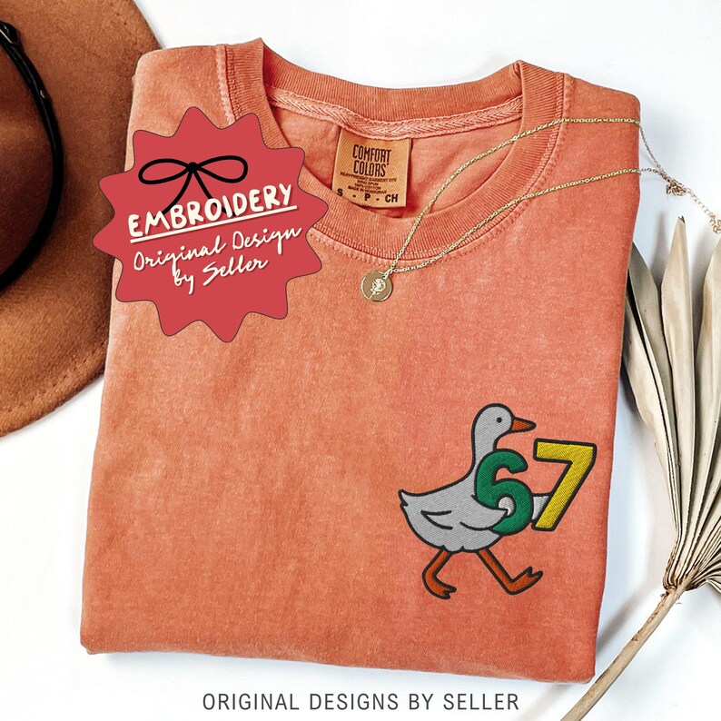 May include: A coral-colored t-shirt with an embroidered design of a goose carrying the number 57. The shirt has a round neckline and short sleeves. A gold necklace with a pendant is draped on the shirt. The text "EMBROIDERY Original Design by Seller" is on a red tag.