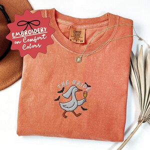 May include: Coral-colored t-shirt with an embroidered design featuring a goose holding a champagne glass, the text "THE BRIDE", and bats. A gold necklace with a pendant is draped on the shirt. The shirt has a Comfort Colors tag.