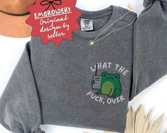 May include: A gray sweatshirt with the embroidered phrase "WHAT THE FUCK, OVER" and a green frog graphic holding a walkie-talkie. The sweatshirt has a crew neck and long sleeves. The text "EMBROIDERY Original design by seller" is also visible.