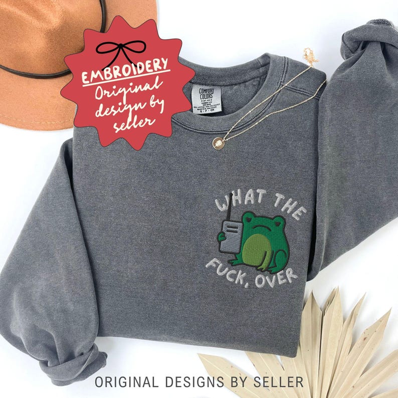 May include: A gray sweatshirt with the embroidered phrase "WHAT THE FUCK, OVER" and a green frog graphic holding a walkie-talkie. The sweatshirt has a crew neck and long sleeves. The text "EMBROIDERY Original design by seller" is also visible.