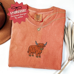 May include: A coral-colored t-shirt with a Highland cow embroidery. The shirt features a crew neck and short sleeves. A gold necklace with a pendant rests on the shirt. The tag reads "Comfort Colors". A red tag with the text "Embroidery Original design by seller" is visible.