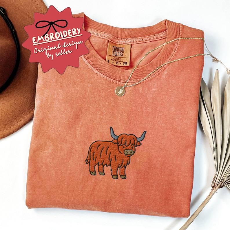 May include: A coral-colored t-shirt with a Highland cow embroidery. The shirt features a crew neck and short sleeves. A gold necklace with a pendant rests on the shirt. The tag reads "Comfort Colors". A red tag with the text "Embroidery Original design by seller" is visible.