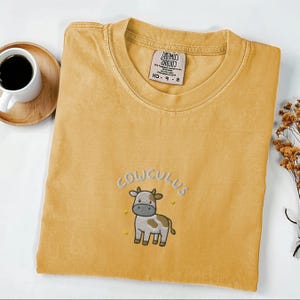May include: Mustard yellow t-shirt with an embroidered cartoon cow and the word "COWCULUS" above it. The shirt has a crew neck and short sleeves. A white coffee mug and a small wooden plate are in the background.