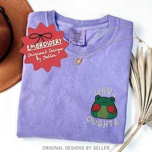 May include: A lilac Comfort Colors t-shirt with an embroidered frog graphic and the words "WHY I OUGHTA". The shirt has a round neckline and short sleeves. A gold necklace with a pendant is draped across the shirt. The text "EMBROIDERY Original Design by Seller" is on a red tag.