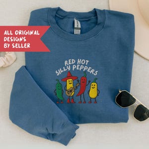 K&ouml;nnte beinhalten: Ein blaues Sweatshirt mit dem gestickten Schriftzug "Red Hot Silly Peppers" und einer Grafik von vier anthropomorphen Chilischoten. Die Chilischoten sind gr&uuml;n, gelb und rot, eine spielt Gitarre. Das Sweatshirt hat einen Rundhalsausschnitt.