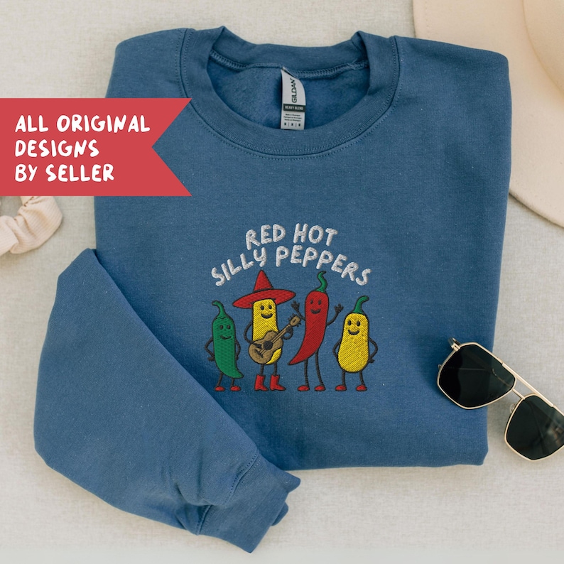 K&ouml;nnte beinhalten: Ein blaues Sweatshirt mit dem gestickten Schriftzug "Red Hot Silly Peppers" und einer Grafik von vier anthropomorphen Chilischoten. Die Chilischoten sind gr&uuml;n, gelb und rot, eine spielt Gitarre. Das Sweatshirt hat einen Rundhalsausschnitt.