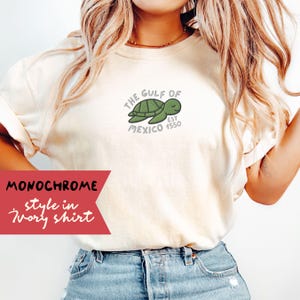 May include: A white t-shirt with a green sea turtle graphic and the text "The Gulf of Mexico Est 1550". The shirt is styled with blue jeans.