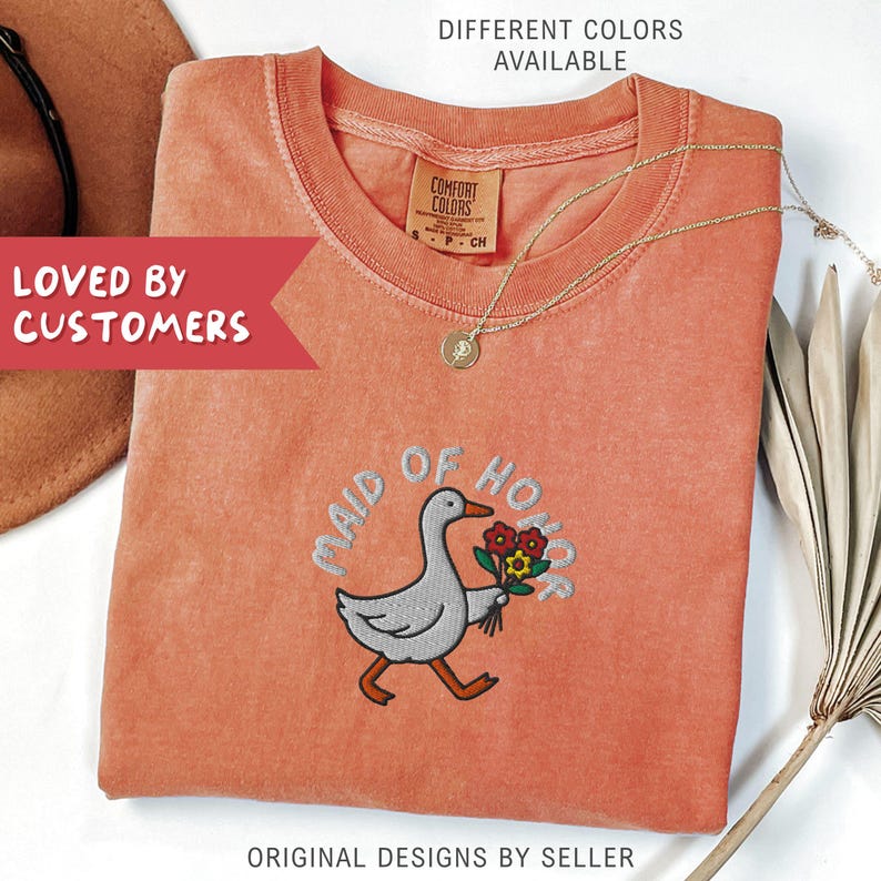 May include: Coral-colored t-shirt with an embroidered design of a goose holding flowers, with the words "MAID OF HONOR" above the goose. A gold necklace and the text "DIFFERENT COLORS AVAILABLE" are also visible.