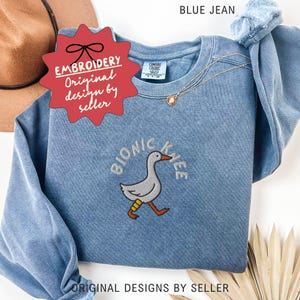 May include: Blue jean sweatshirt with an embroidered design of a goose with a bionic knee. The text "BIONIC KNEE" arches above the goose. The sweatshirt has long sleeves and a crew neck. The text "BLUE JEAN" is at the top.