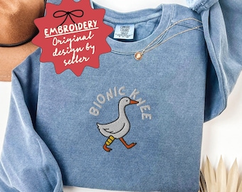 May include: Blue jean sweatshirt with an embroidered design of a goose with a bionic knee. The text "BIONIC KNEE" arches above the goose. The sweatshirt has long sleeves and a crew neck. The text "BLUE JEAN" is at the top.