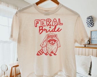 Może przedstawiać: Kremowa koszulka z czerwonym napisem "FERAL Bride", nad czerwoną ilustracją szopa w welonie. Koszulka wisi na drewnianym wieszaku. Tekst "ORIGINAL DESIGNS BY SELLER" znajduje się na dole.