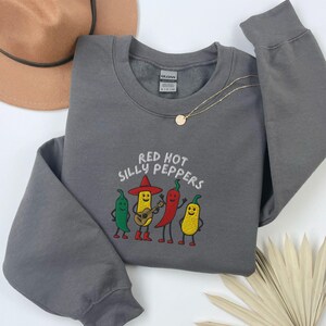 K&ouml;nnte beinhalten: Ein graues Sweatshirt mit dem gestickten Text "Red Hot Silly Peppers" &uuml;ber einer Grafik von vier anthropomorphen Chilischoten. Eine Chilisorte spielt Gitarre. Eine goldene Halskette und ein brauner Hut sind ebenfalls sichtbar.