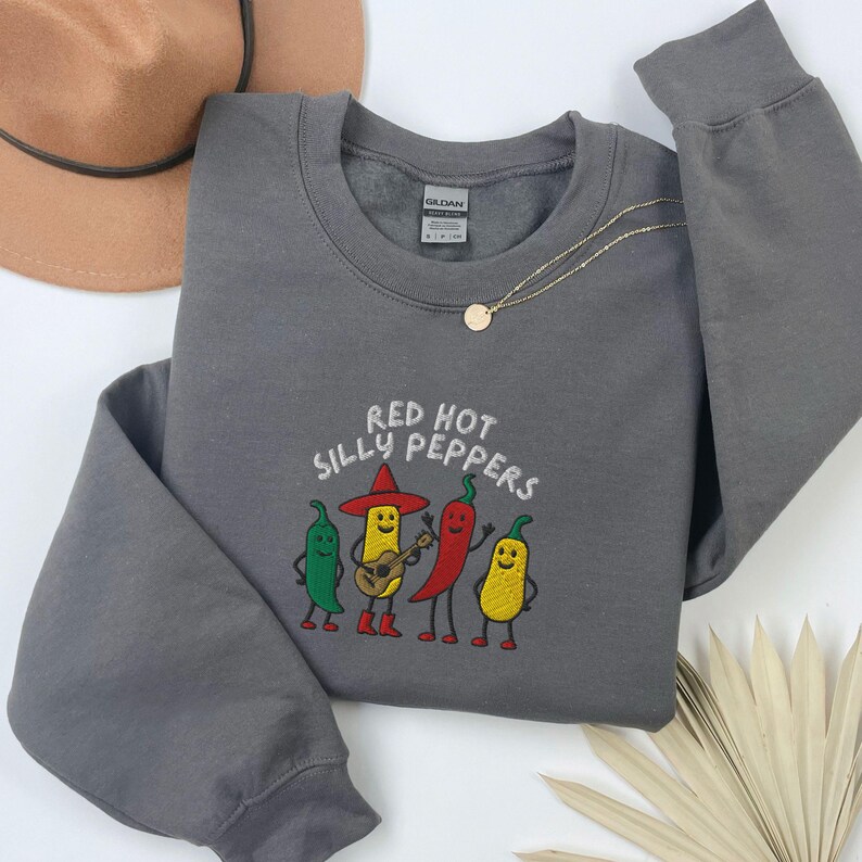 K&ouml;nnte beinhalten: Ein graues Sweatshirt mit dem gestickten Text "Red Hot Silly Peppers" &uuml;ber einer Grafik von vier anthropomorphen Chilischoten. Eine Chilisorte spielt Gitarre. Eine goldene Halskette und ein brauner Hut sind ebenfalls sichtbar.