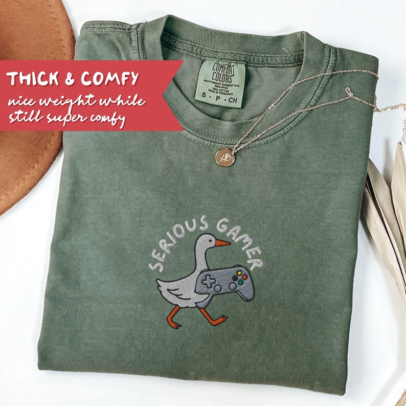 May include: A sage green t-shirt with the embroidered phrase "SERIOUS GAMER" and a cartoon goose holding a video game controller. The shirt also features the text "THICK & COMFY nice weight while still super comfy". A gold necklace with a pendant is draped on the shirt.