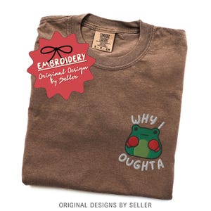 May include: Brown t-shirt with a crew neck and short sleeves. The shirt features an embroidered frog graphic with the text "WHY I OUGHTA". A red starburst sticker with the word "EMBROIDERY" is on the shirt.