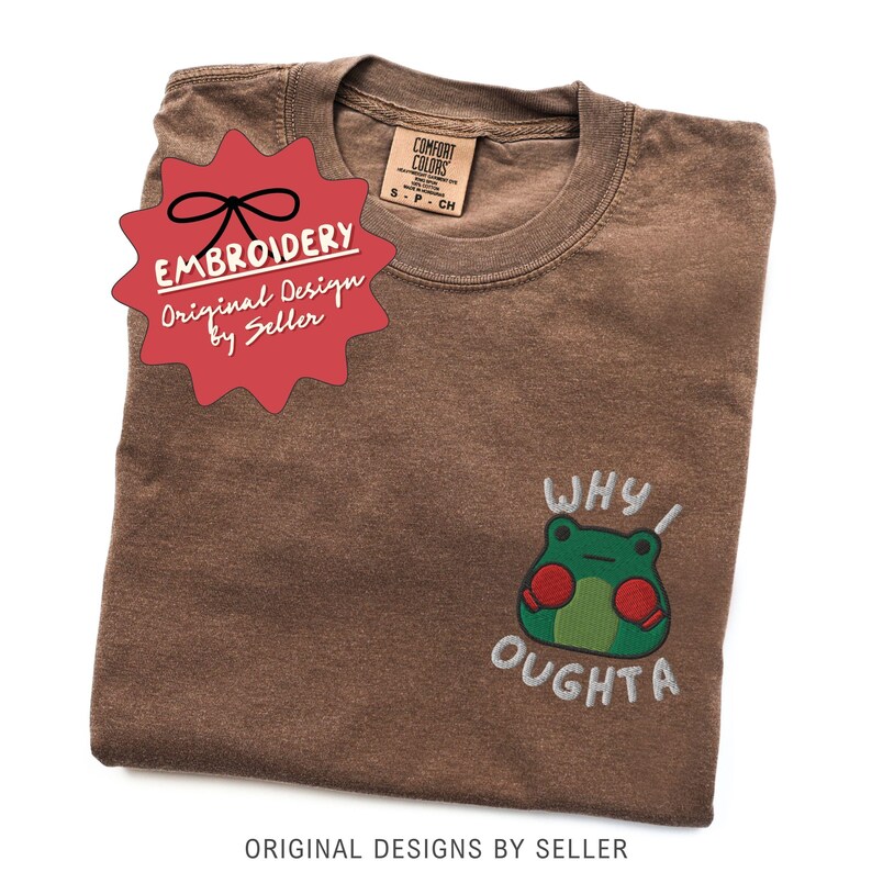 May include: Brown t-shirt with a crew neck and short sleeves. The shirt features an embroidered frog graphic with the text "WHY I OUGHTA". A red starburst sticker with the word "EMBROIDERY" is on the shirt.