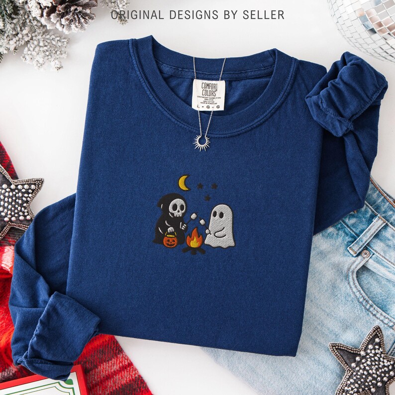 May include: A navy blue long-sleeved shirt with an embroidered Halloween design. The design features a skeleton, a ghost, a campfire, a moon, and stars. The shirt has a crew neck and a Comfort Colors tag. The text "ORIGINAL DESIGNS BY SELLER" is visible.