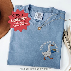 May include: A blue Comfort Colors t-shirt with an embroidered design of a goose carrying a bowl, with the text "TOTALLY KILN IT". A gold necklace and a red tag that says "EMBROIDERY on Comfort Colors Shirts" are also visible.