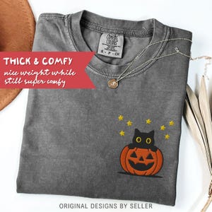 Może przedstawiać: Szara koszulka Comfort Colors z haftowanym wzorem czarnego kota w pomarańczowej dyni, otoczonego ż&oacute;łtymi gwiazdami. Koszulka ma napis "THICK & COMFY" i "ORIGINAL DESIGNS BY SELLER". Srebrny naszyjnik jest ułożony na koszulce.