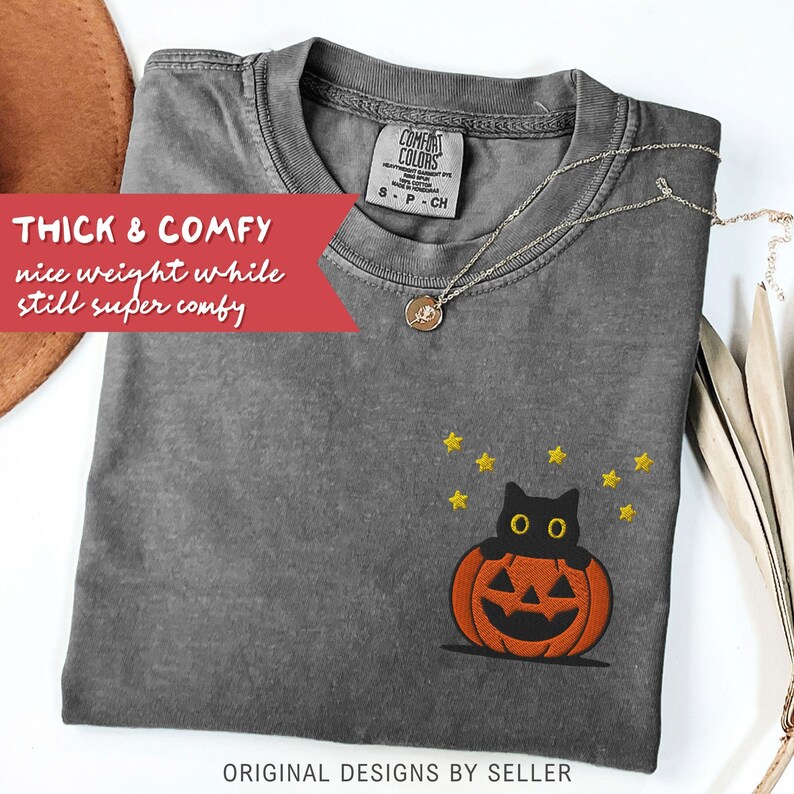 Może przedstawiać: Szara koszulka Comfort Colors z haftowanym wzorem czarnego kota w pomarańczowej dyni, otoczonego ż&oacute;łtymi gwiazdami. Koszulka ma napis "THICK & COMFY" i "ORIGINAL DESIGNS BY SELLER". Srebrny naszyjnik jest ułożony na koszulce.