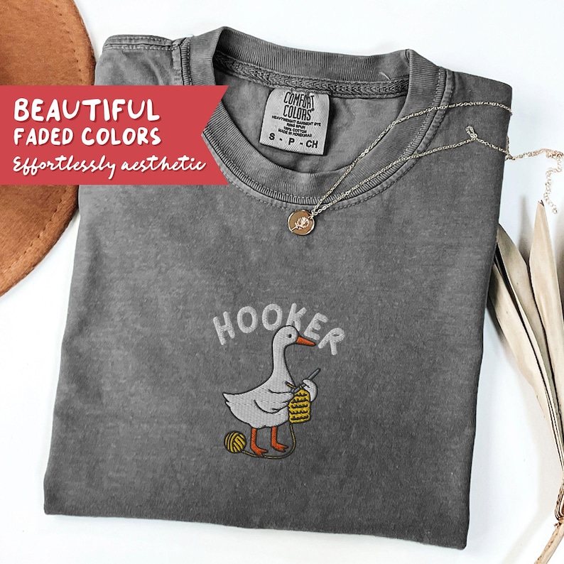 May include: Gray Comfort Colors t-shirt with the word "Hooker" above a cartoon goose knitting. The goose is holding a crochet hook and yarn. The shirt has a necklace and the text "BEAUTIFUL FADED COLORS Effortlessly aesthetic".