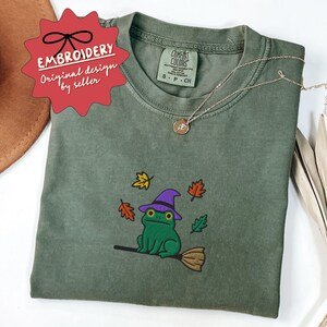 May include: Olive green t-shirt with an embroidered design of a green frog wearing a purple witch hat and riding a broom, surrounded by autumn leaves. The shirt has the text "Comfort Colors" on the tag. A gold necklace is draped across the shirt.