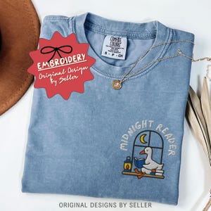 May include: A blue Comfort Colors t-shirt with an embroidered design featuring a duck reading a book by a window, with the words "MIDNIGHT READER". A gold necklace is draped on the shirt. The shirt has the text "ORIGINAL DESIGNS BY SELLER".