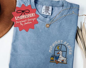 Peut inclure: Un t-shirt bleu Comfort Colors avec un motif brodé d'un canard lisant près d'une fenêtre, avec les mots "MIDNIGHT READER". Un collier en or est posé sur le t-shirt. Le t-shirt porte l'inscription "ORIGINAL DESIGNS BY SELLER".