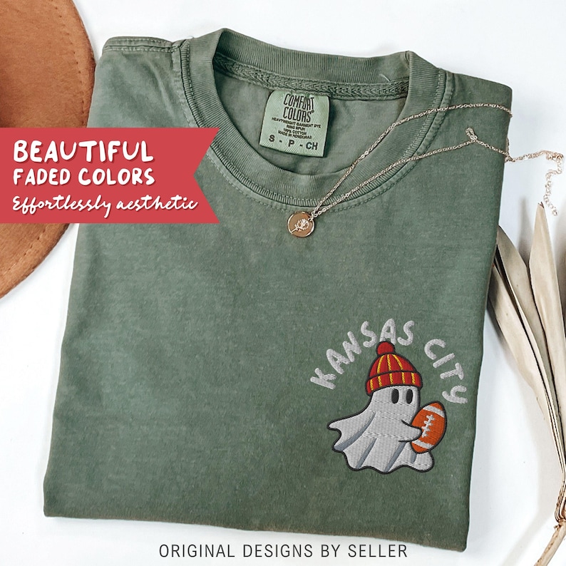 May include: A sage green t-shirt with an embroidered design of a ghost wearing a red and yellow beanie and holding a football. The text "Kansas City" is above the ghost. The shirt also has the text "BEAUTIFUL FADED COLORS Effortlessly aesthetic" and "ORIGINAL DESIGNS BY SELLER".