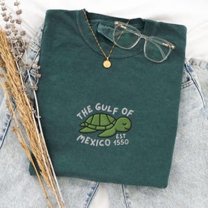 May include: A dark green t-shirt with a white embroidered sea turtle and the text "The Gulf of Mexico Est 1550".