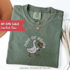 May include: Olive green t-shirt with the embroidered phrase "MAID OF HONOR" and a cartoon goose holding flowers. A gold necklace and a brown hat are also visible. The shirt has a "COMFORT COLORS" tag. Text on the image reads "40-50% SALE Limited Time".