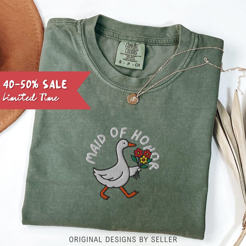 May include: Olive green t-shirt with the embroidered phrase "MAID OF HONOR" and a cartoon goose holding flowers. A gold necklace and a brown hat are also visible. The shirt has a "COMFORT COLORS" tag. Text on the image reads "40-50% SALE Limited Time".