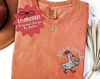 May include: Coral-coloured t-shirt with an embroidered design of a duck in a Santa hat, holding a coffee cup, and the words "SILLY JUICE". A gold necklace and a red tag that says "EMBROIDERY Original Design by Seller" are also visible.