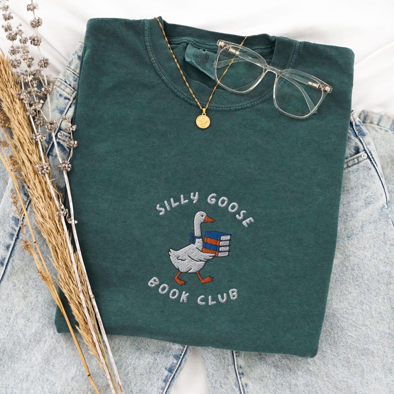 Może przedstawiać: Zielony T-shirt z haftowanym wzorem gęsi niosącej stos książek. Tekst "Silly Goose Book Club" jest wyszyty wok&oacute;ł gęsi.
