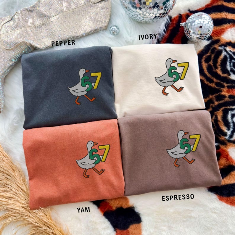 May include: Four folded t-shirts in pepper, ivory, yam, and espresso colors. Each shirt features an embroidered design of a walking duck carrying the number 7 in green and yellow. The shirts are displayed on a patterned surface.