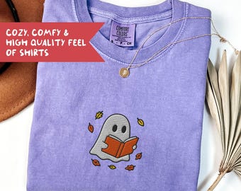 May include: A lavender Comfort Colors t-shirt with an embroidered ghost reading a book, surrounded by autumn leaves. The shirt has the text "COZY, COMFY & HIGH QUALITY FEEL OF SHIRTS" on a red banner. A gold necklace with a pendant is draped on the shirt.