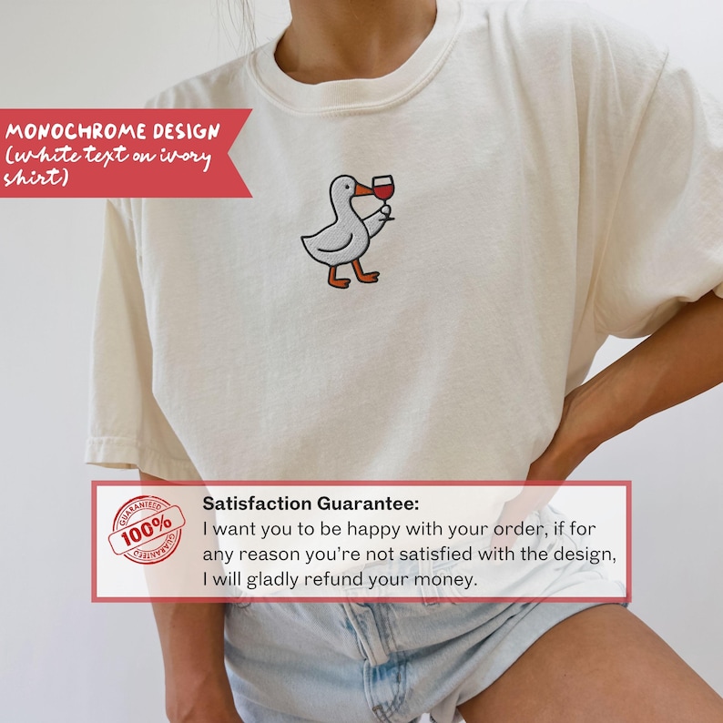 May include: Ivory-colored t-shirt featuring a cartoon duck holding a wine glass. The duck is white with orange feet and a black outline. The shirt has the text "MONOCHROME DESIGN" in white. The shirt is made of a soft material.