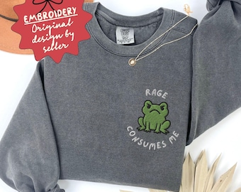 May include: A gray sweatshirt with a green frog embroidery that says "Rage Consumes Me." The sweatshirt has long sleeves and a crew neck. The image also includes a gold necklace and a red tag that says "Embroidery Original design by seller."