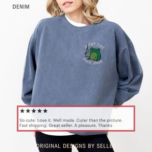 May include: Denim blue sweatshirt with the text "WHAT THE FUCK, OVER" and a cartoon frog holding a coffee cup. The sweatshirt has a crew neck and long sleeves. The word "DENIM" is printed at the top left.